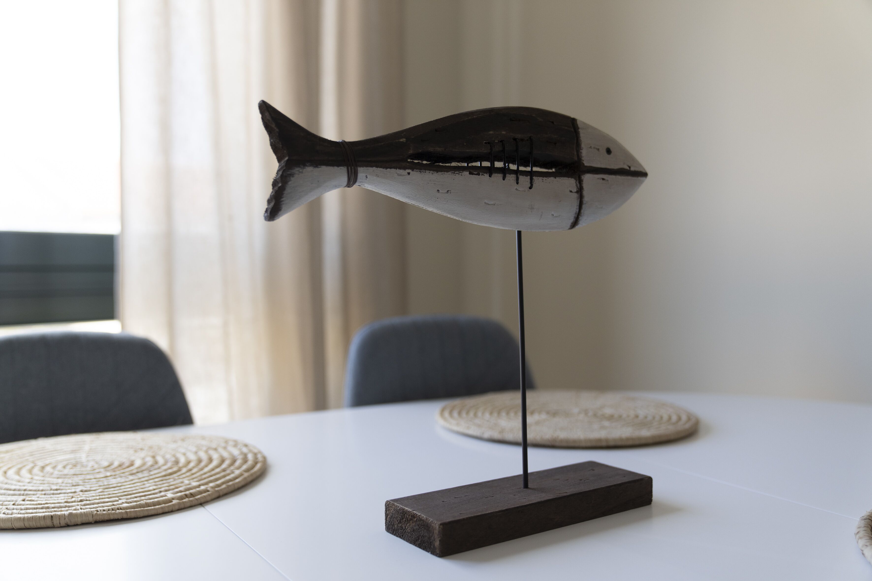 A table with a fish-shaped decoration on a stand, two woven placemats, and a chair in the background.