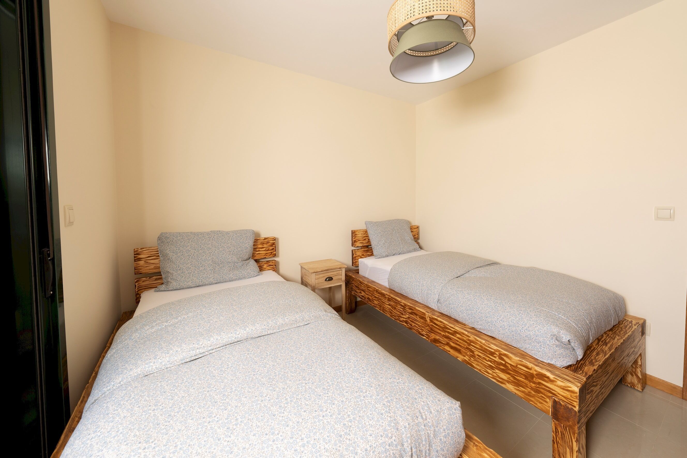 Une chambre avec deux lits en bois, une table de chevet et un luminaire suspendu au plafond, fait de fibres tressées.