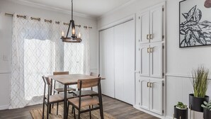 Comedor con mesa y sillas de madera, candelabro, paredes blancas y un amplio ventanal con cortinas estampadas.