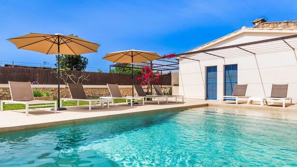 Una piscina con sillones de descanso y sombrillas, un muro de piedra y un edificio blanco con contraventanas azules.