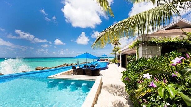 A pool area with a view of the ocean, outdoor seating, and tropical plants.