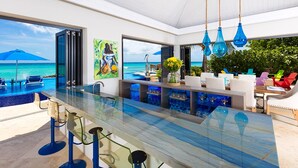 A modern kitchen with a glass countertop, bar area, and seating by a pool with a view of the ocean.