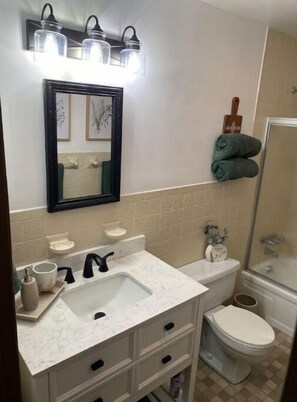 Casa de banho com uma pia branca, um espelho e uma sanita. Toalhas penduradas na parede e zona de duche.
