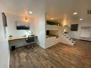 Quarto moderno e compacto, com piso de madeira, uma escrivaninha com cadeira, uma cama com escada e televisão na parede.