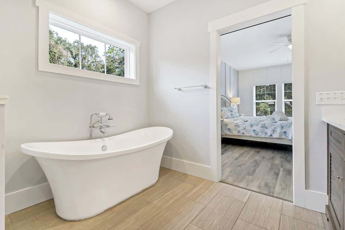 A modern bathroom with a freestanding bathtub, a window, and a view into a bedroom with a bed and a nightstand.