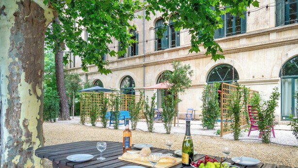An outdoor dining area with a table set for a meal, surrounded by trees and a building with arched windows. - Aura - Apartment for 8 persons in renovated Couvent les Ursulines, Pézenas (Pézenas)