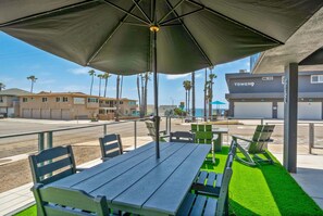 An outdoor seating area with tables and chairs, a large umbrella, and a view of the beach and buildings in the background.