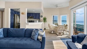 A modern living room with a blue sofa, a dining area with wicker chairs, a kitchen with blue cabinets, and a view of the ocean. - Beachfront Luxury 2BR with Ocean-View Balconies & Spectacular Sunsets (Oceanside)
