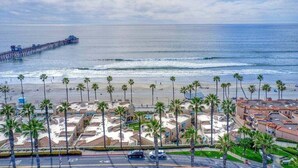 A coastal hotel with a beach view, palm trees, and a pier in the background. - Beachfront Luxury 2BR with Ocean-View Balconies & Spectacular Sunsets (Oceanside)