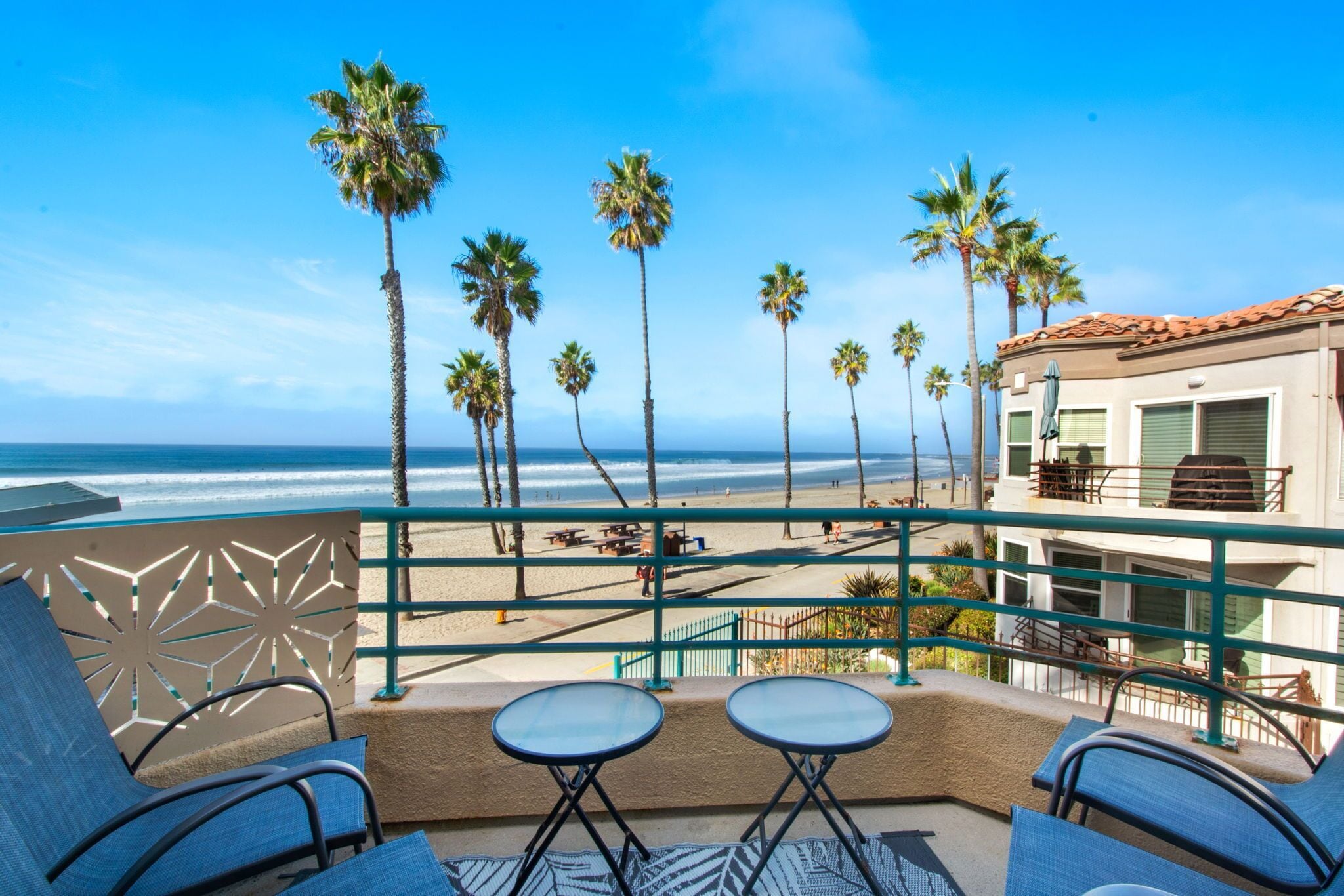 A balcony with two tables and chairs, overlooking a beach with palm trees and a building with a red roof.