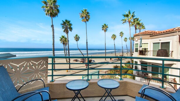 A balcony with two tables and chairs, overlooking a beach with palm trees and a building with a red roof. - Beachfront Luxury 2BR with Ocean-View Balconies & Spectacular Sunsets (Oceanside)