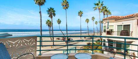 A balcony with two tables and chairs, overlooking a beach with palm trees and a building with a red roof.