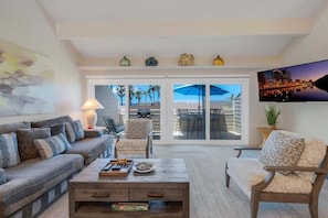 A living room with a sofa, armchairs, a coffee table, and a TV. There is a sliding glass door leading to an outdoor patio with a table and chairs. - Seabluffe Coastal Retreat, 3BD Facing Grandview Surf Beach, Ocean Views, Pools, Spa (Encinitas)