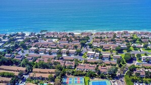 Vue aérienne d’un quartier résidentiel situé sur la côte, avec des maisons, un court de tennis et l’océan.