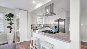 A modern kitchen with white cabinetry, a central island with bar stools, a stainless steel refrigerator, and a large window with blinds.