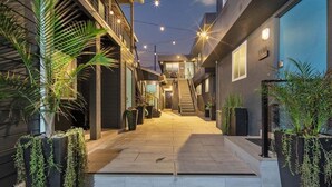 A modern building with a staircase leading to an upper level, surrounded by potted plants and string lights. - Oceanview Escape | Tower 9 Condo Steps from the Beach (Oceanside)
