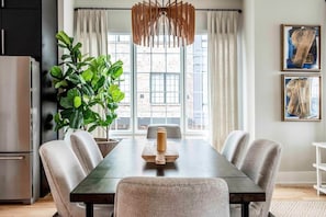 Uma sala de jantar com uma mesa comprida, seis cadeiras, um vaso de planta, um centro de mesa de madeira e uma luminária pendente moderna.