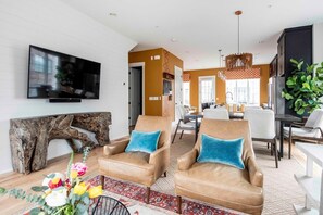 A modern living room with leather armchairs, a flat-screen TV, a wooden art piece, and a dining area with a chandelier.