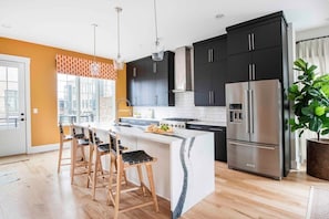 Uma cozinha moderna com ilha central, armários pretos, eletrodomésticos de aço inoxidável e uma janela grande com cortinas laranja.