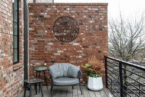 A balcony with a brick wall, a round metal artwork, a grey armchair, a small table, and a potted plant.