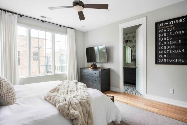 A bedroom with a bed, a ceiling fan, a dresser, a television, and a wall with a departures sign listing various cities.