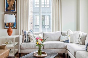 A modern living room with a light-colored sofa, a coffee table with a vase of flowers, and a large window with curtains.