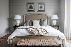 A bedroom with a large bed, two bedside tables with lamps, a bench at the foot of the bed, and two framed pictures on the wall.