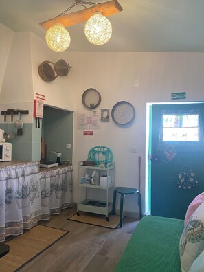 Una cocina con microondas, tostadora y un estante pequeño con utensilios. Hay una puerta con un cartel encima, un espejo y un ventilador de pared.