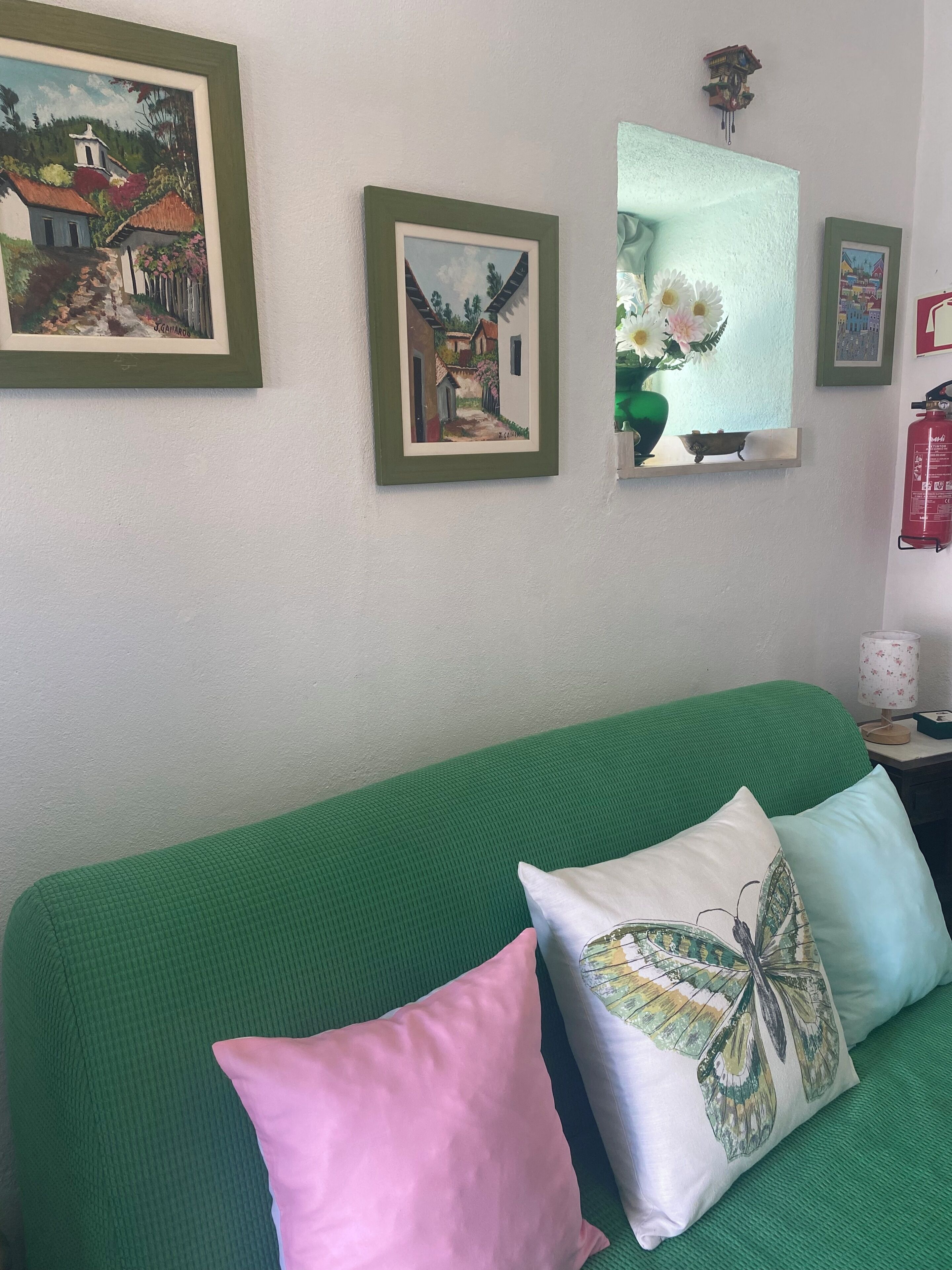 Un sofá verde con dos almohadas, un cuadro enmarcado, un jarrón con flores y un extintor en la pared.