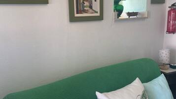Un sofá verde con dos almohadas, un cuadro enmarcado, un jarrón con flores y un extintor en la pared.