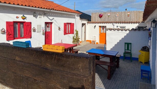 A white building with red shutters, a wooden bench, and a table with chairs.