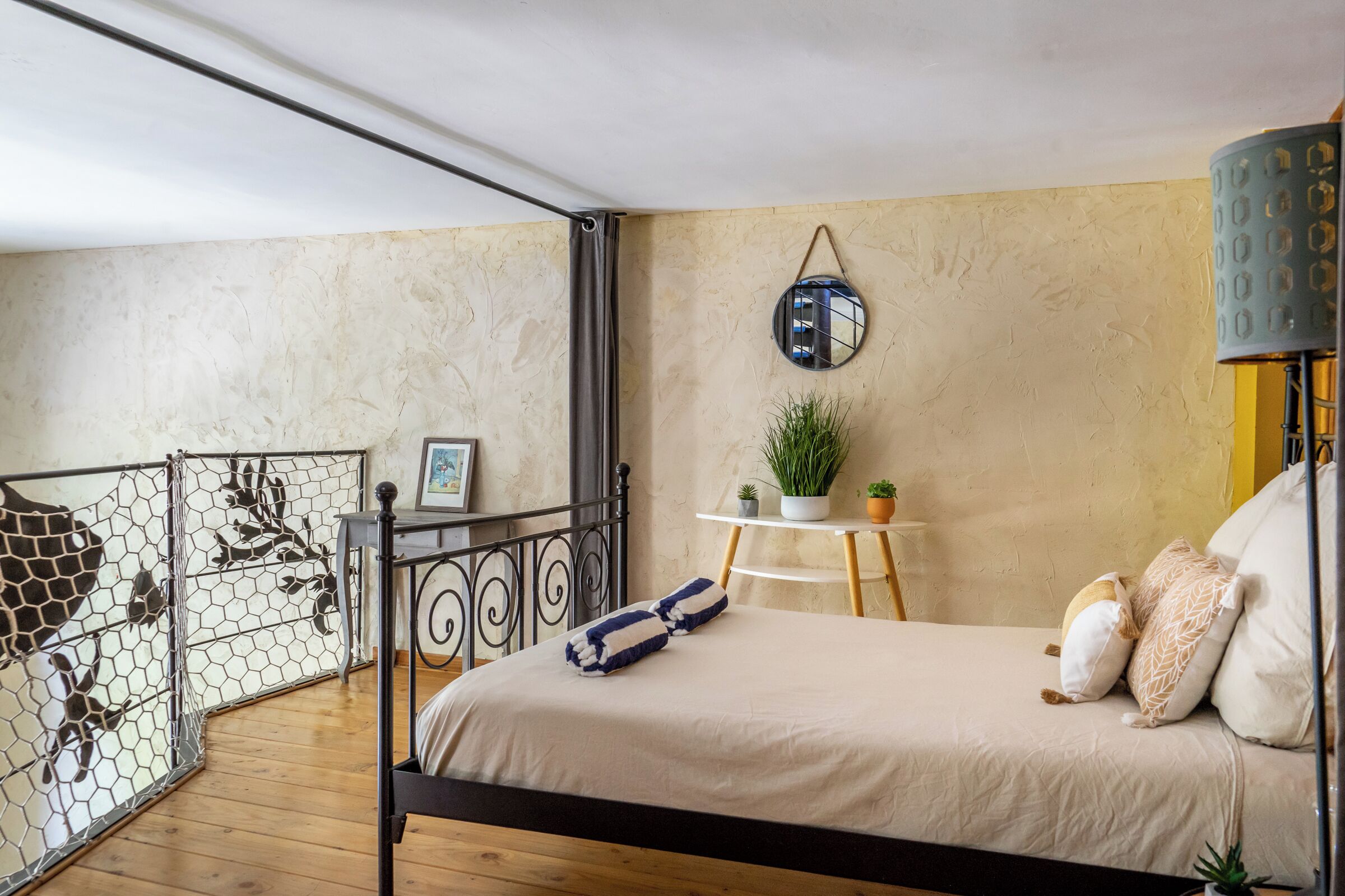 Een slaapkamer met een bed, een klein tafeltje met planten, een hangspiegel en uitzicht op een balkon met een metalen hek.