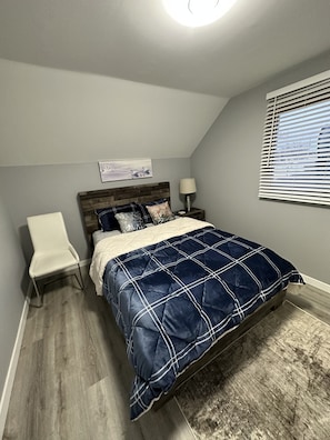 Une chambre à coucher comprenant un lit, une chaise, une table de chevet, une lampe, une fenêtre avec des stores et un tableau accroché au mur.