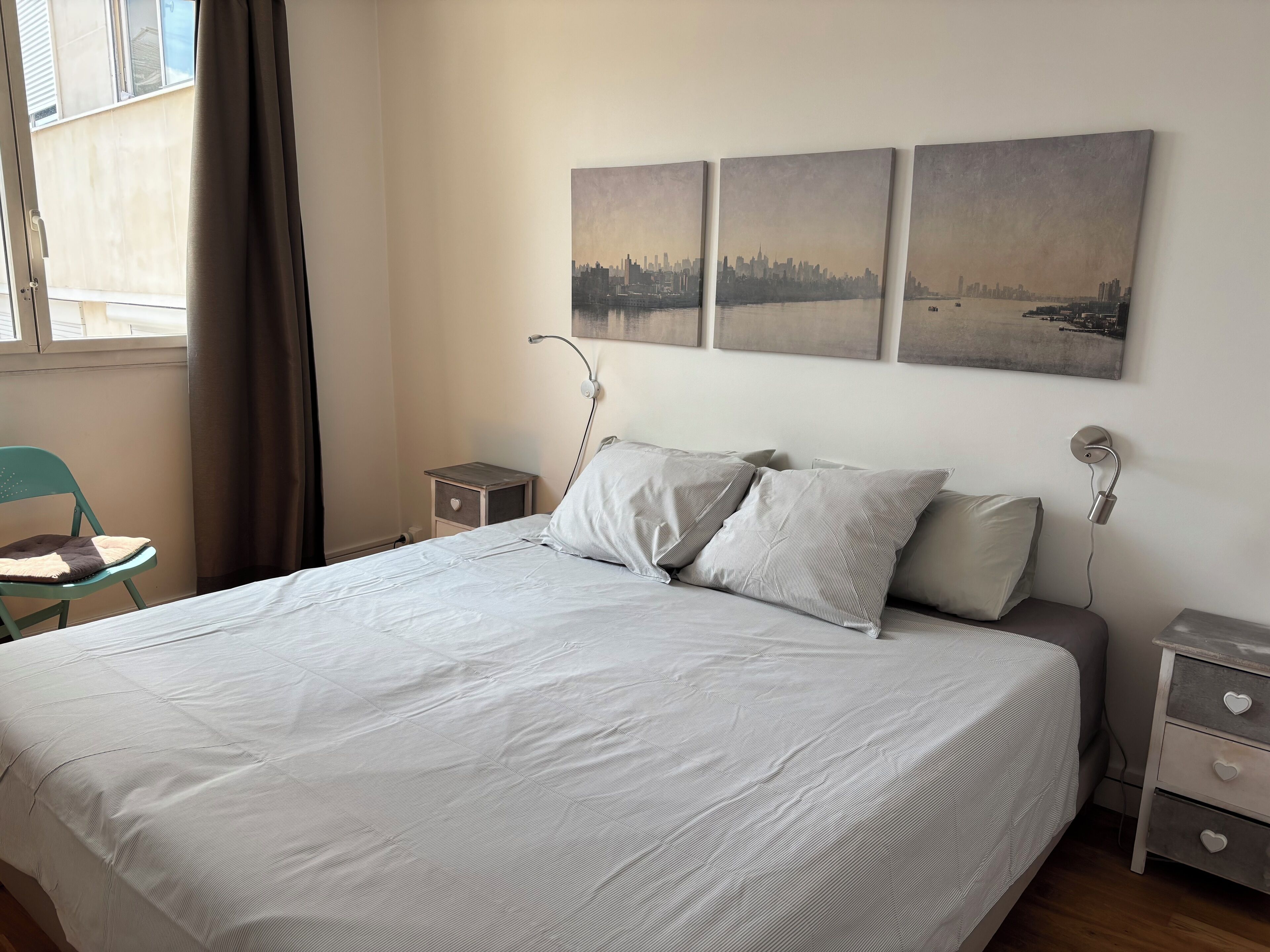 A neatly made bed with a white bedspread, two pillows, and a nightstand with a drawer. There are three wall-mounted paintings above the bed.