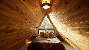 A cozy, triangular wooden room with a bed, a window, and a light fixture. - Elwood's A-frame · Modern A-frame w/ hot tub + view (Florissant)
