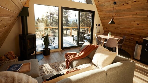 A cozy living room with a sofa, a coffee table, and a fireplace. There is a large sliding glass door leading to an outdoor deck. - Elwood's A-frame · Modern A-frame w/ hot tub + view (Florissant)