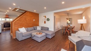 Ein modernes Wohnzimmer mit einem grauen Ecksofa, einer Holztreppe, einem Essbereich mit Holzsesseln und einer Küche mit Backofen und Herd.