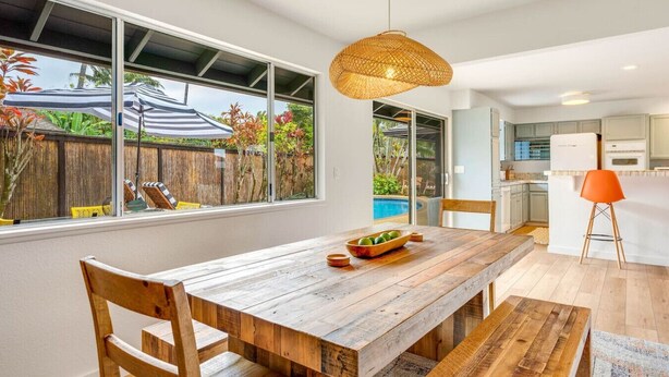 A modern kitchen with white cabinets, a wooden dining table, and a large window overlooking a backyard with a pool. - Cool, Lo-Fi Styled Surf Shack - Ka Hale Nalu (Princeville)