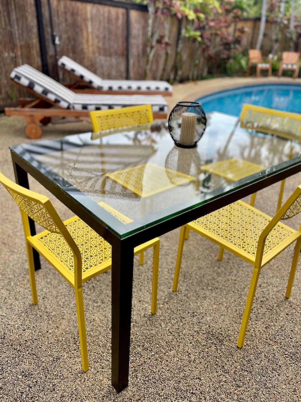 A glass-top table with four yellow chairs, a small decorative vase, and a pool in the background.