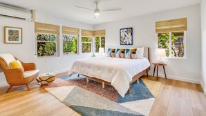 A bedroom with a large bed, a chair, a small table, and a window with a view of greenery. - Cool, Lo-Fi Styled Surf Shack - Ka Hale Nalu (Princeville)