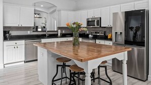 Cocina moderna con armarios blancos, electrodomésticos de acero inoxidable, una isla de madera con taburetes y un jarrón con flores.