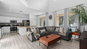 Un amplio salón con un sofá seccional gris, una mesa de centro de madera y una cocina al fondo.