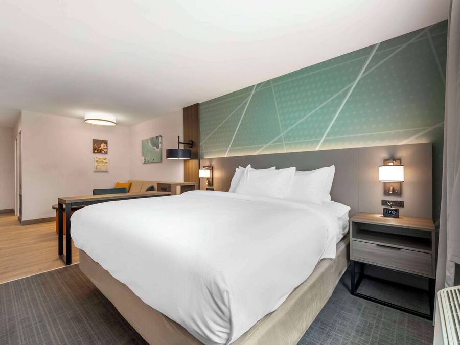 A hotel room with a large bed, a desk, a chair, and a nightstand with a lamp.