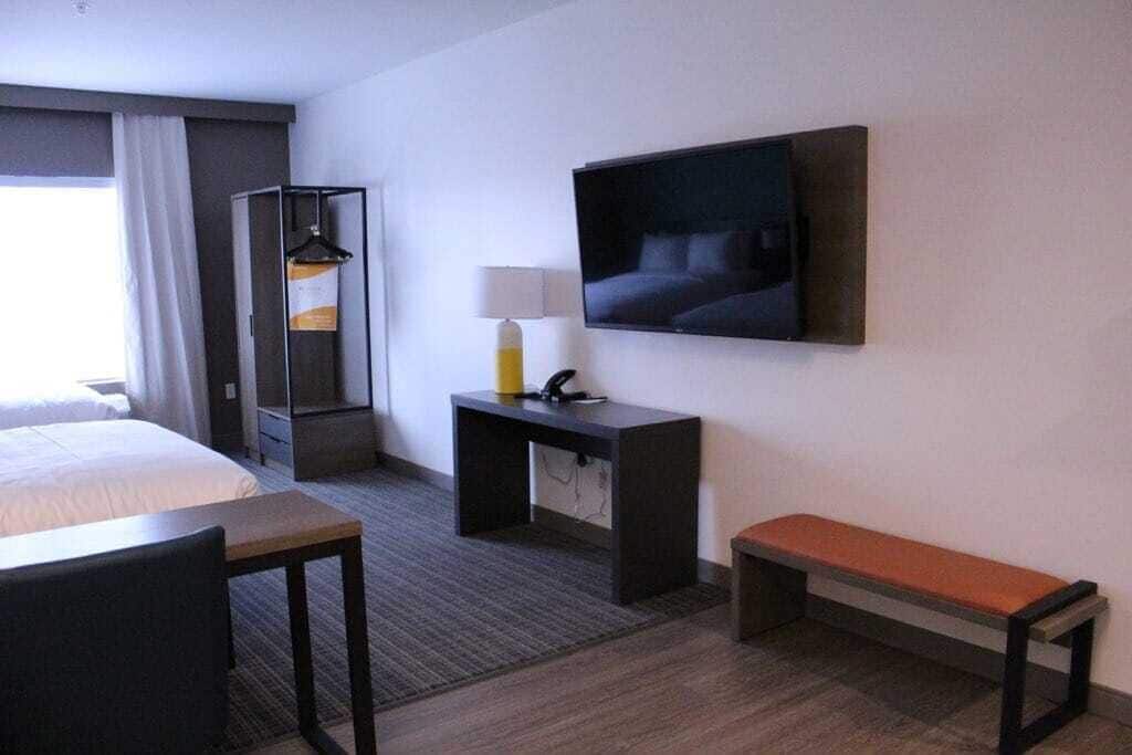 A hotel room with a bed, a desk, a bench, a TV, a coat rack, and a lamp.