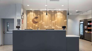 A modern reception desk with a wooden panel backdrop, a crescent-shaped light fixture, and a refrigerator stocked with beverages. - Comfort Suites Richmond East (Richmond)