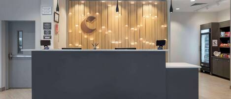 Un mostrador de recepción moderno con un panel de madera de fondo, una lámpara en forma de luna creciente y un refrigerador lleno de bebidas.