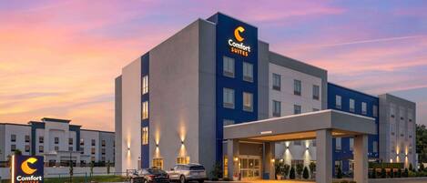 Un hotel Comfort Suites con diseño moderno, destacando una paleta de colores azul y blanco, una entrada bien iluminada y un estacionamiento con varios autos.