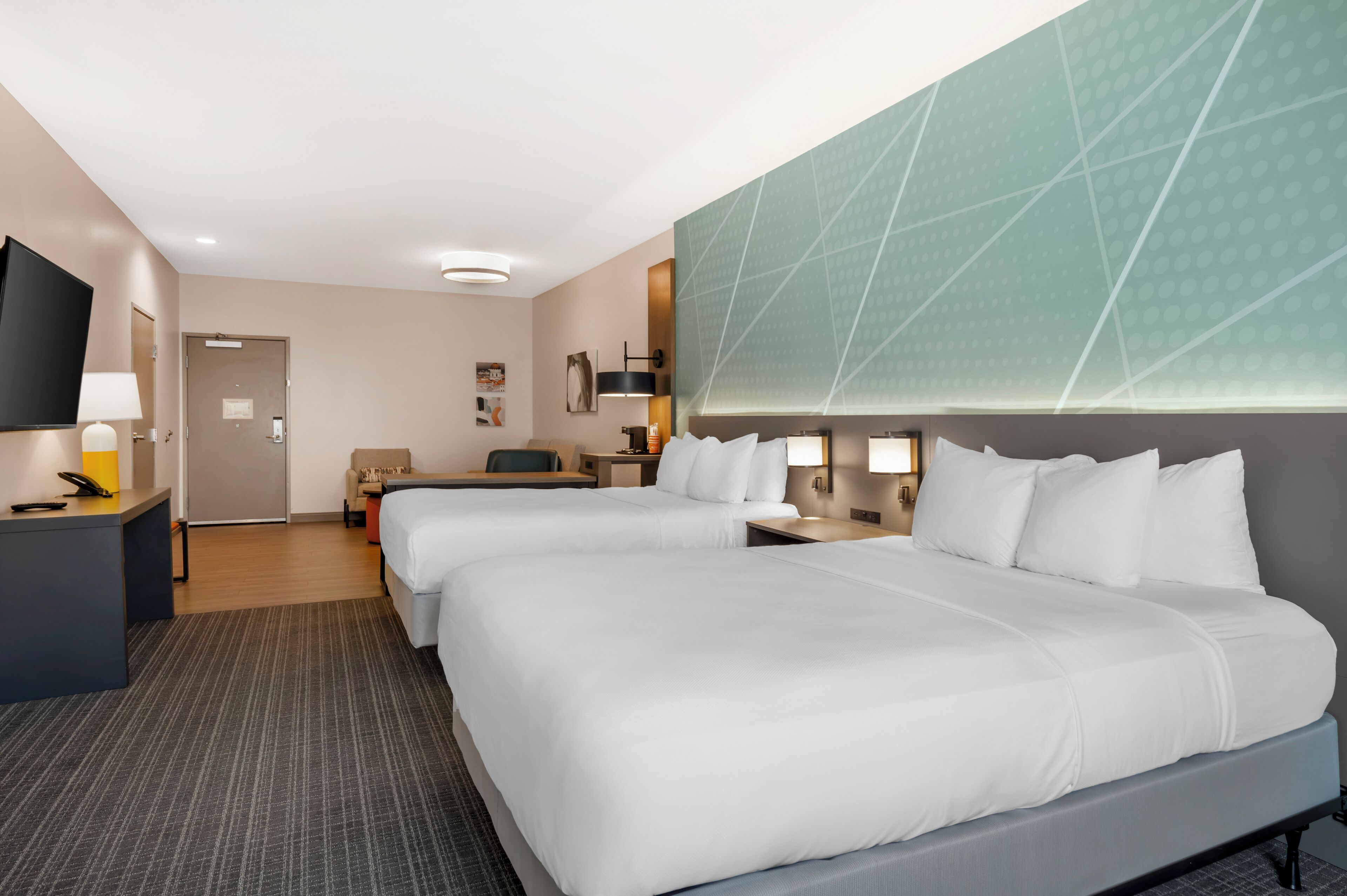A hotel room with two beds, a desk with a lamp, a TV, and a wall with a geometric pattern.