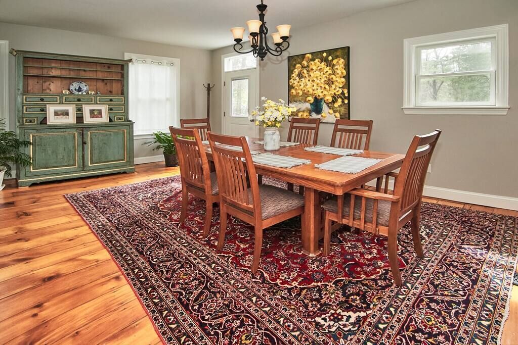 Une salle à manger avec une table en bois, six chaises, un tapis à motifs, un lustre et un buffet vert orné de tableaux encadrés.