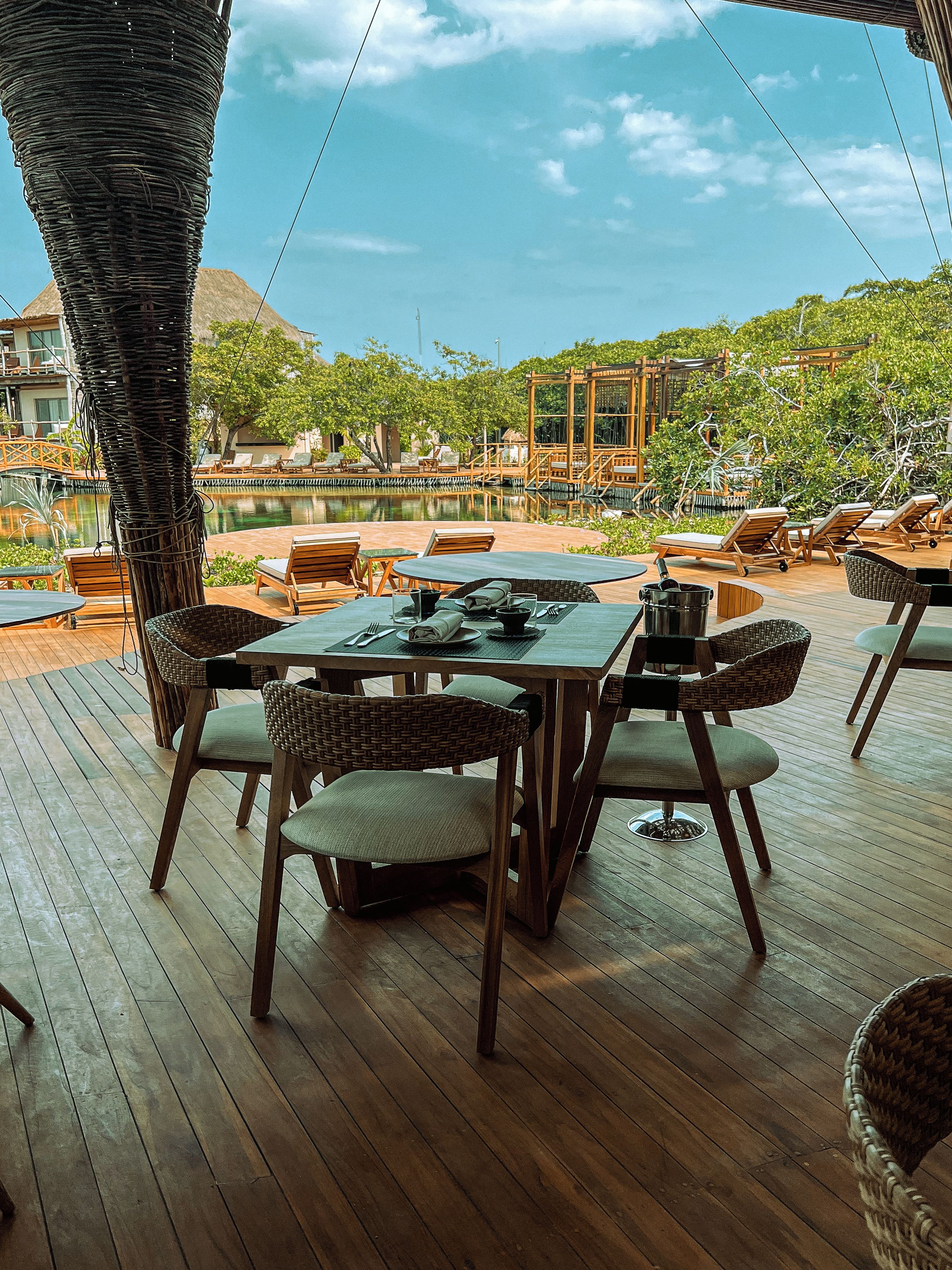 Un espace extérieur aménagé pour les repas, avec des tables et des chaises en bois, une piscine et une vue sur un paysage tropical.